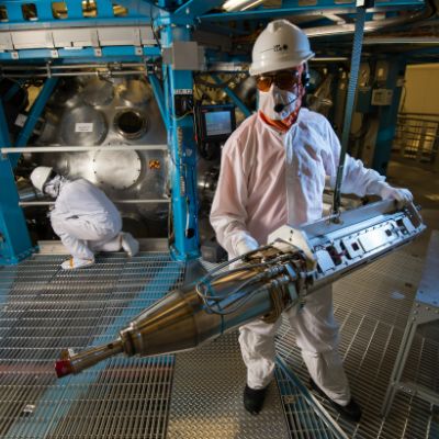 A Laser Lab employee holds a large instrument outside of a target chamber, while wearing a hard hat and protective lab gear.