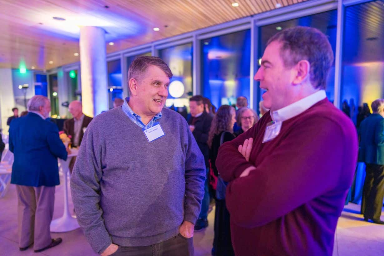 two men in sweaters standing and speaking at a reception