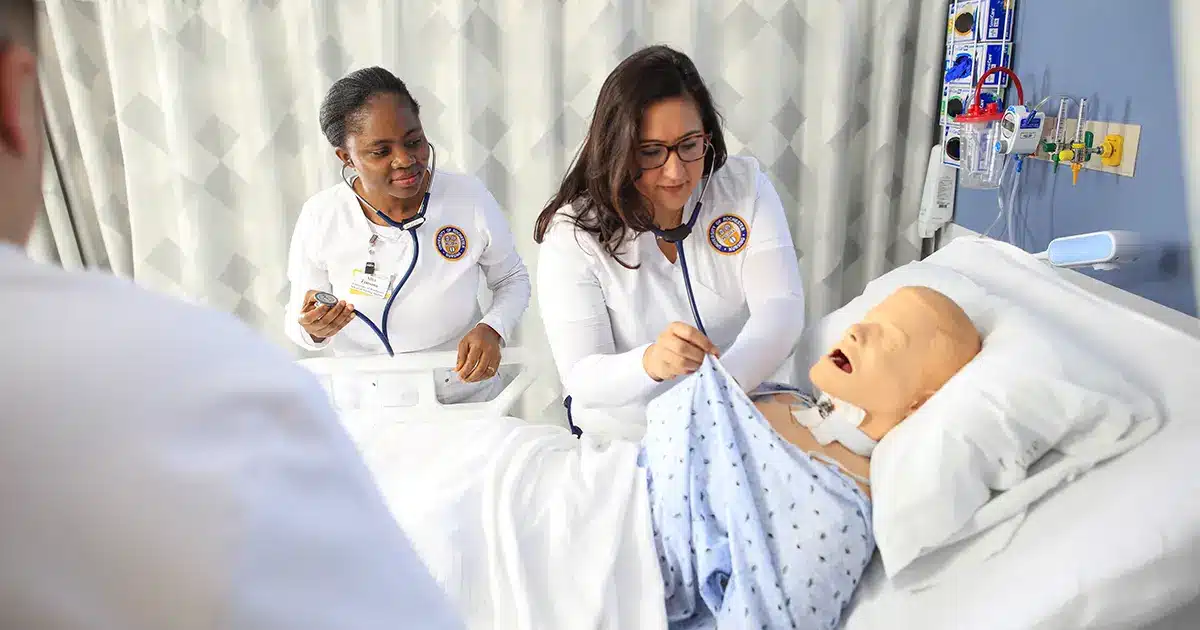 Medical training with two people in scrubs interacting with a mannequin on a hospital bed.